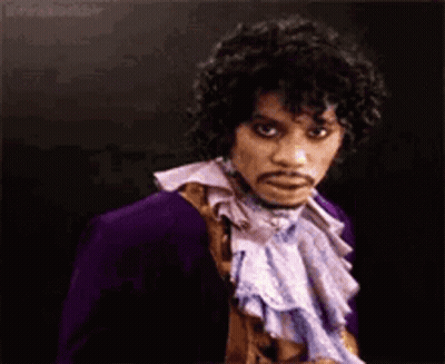 A Man With Curly Hair And A Mustache Is Wearing A Purple Jacket And A Purple Scarf . GIF