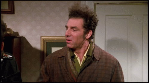 A Man With Curly Hair And A Plaid Jacket Stands In Front Of A Door GIF