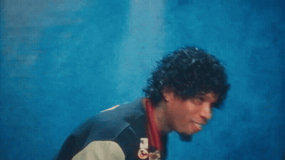 A Man With Curly Hair And A Varsity Jacket Is Standing In Front Of A Blue Wall . GIF
