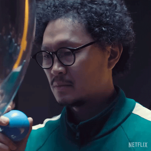 A Man With Curly Hair And Glasses Is Holding A Blue Ball In His Right Hand . GIF