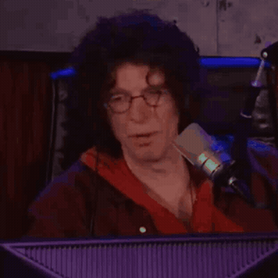 A Man With Curly Hair And Glasses Is Sitting In Front Of A Microphone In Front Of A Computer Monitor . GIF