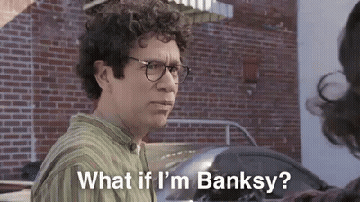 A Man With Curly Hair And Glasses Is Standing In Front Of A Brick Wall And Talking To A Woman . GIF