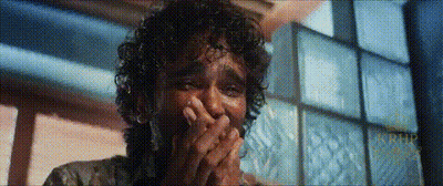 A Man With Curly Hair Is Covering His Mouth With His Hands In A Room . GIF