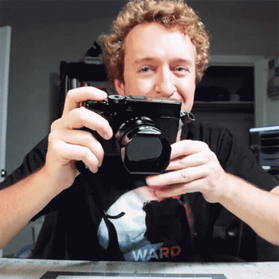 A Man With Curly Hair Is Holding A Camera And Wearing A Ward Shirt GIF