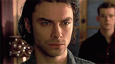 A Man With Curly Hair Is Looking At The Camera While Another Man Stands Behind Him . GIF