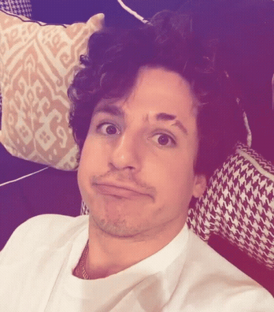 A Man With Curly Hair Is Making A Funny Face In Front Of A Patterned Pillow GIF