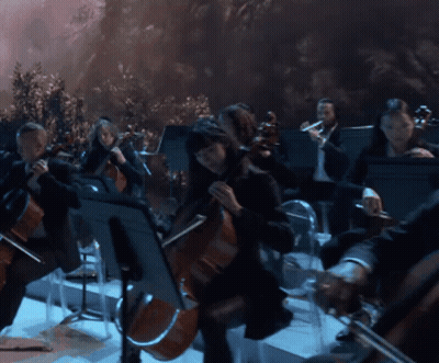 A Man With Curly Hair Is Playing A Flute In Front Of An Orchestra GIF