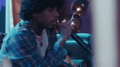 A Man With Curly Hair Is Singing Into A Microphone While Sitting On A Couch GIF