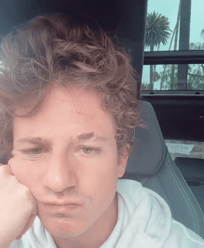 A Man With Curly Hair Is Sitting In A Car With His Hand To His Face GIF