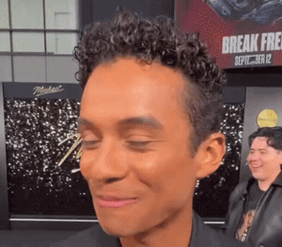 A Man With Curly Hair Is Smiling With His Eyes Closed In Front Of A Sign That Says Break Free GIF