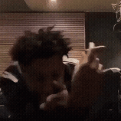 A Man With Curly Hair Is Smoking A Cigarette In Front Of A Wooden Wall . GIF