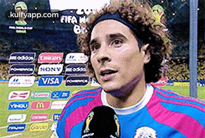 A Man With Curly Hair Is Talking Into A Microphone On A Soccer Field GIF