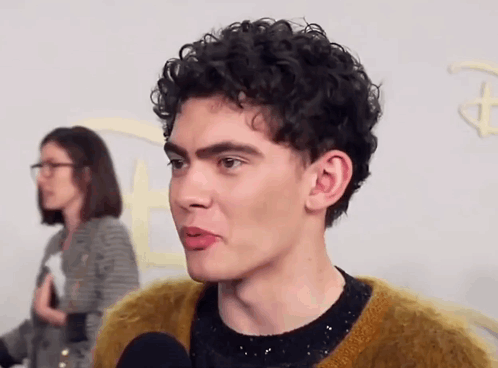 A Man With Curly Hair Is Talking Into A Microphone While A Woman Stands Behind Him . GIF