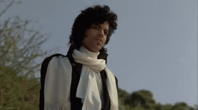 A Man With Curly Hair Is Wearing A White Scarf Around His Neck And A Black Jacket . GIF