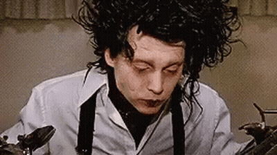 A Man With Curly Hair Is Wearing A White Shirt And Suspenders And Holding A Pair Of Scissors . GIF