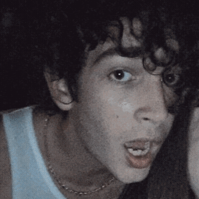 A Man With Curly Hair Is Wearing A White Tank Top And A Necklace GIF