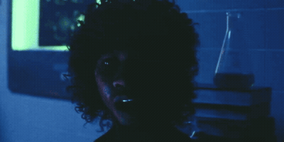 A Man With Curly Hair Looks At The Camera In A Dark Room GIF