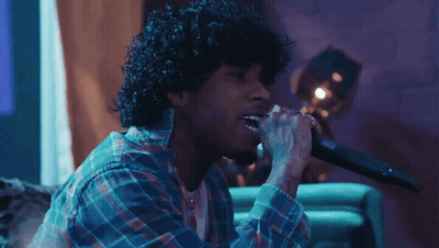 A Man With Curly Hair Singing Into A Microphone While Wearing A Plaid Shirt GIF