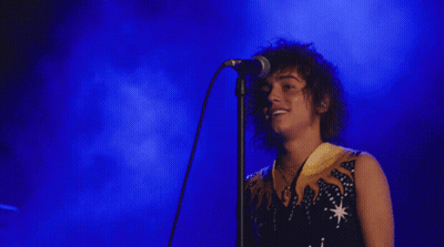 A Man With Curly Hair Singing Into A Microphone With A Blue Background GIF