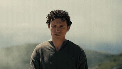 A Man With Curly Hair Wearing A Grey Shirt Stands In Front Of A Mountain GIF