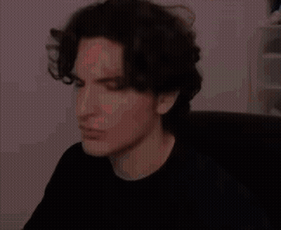 A Man With Dark Hair Is Sitting In A Chair And Making A Funny Face . GIF