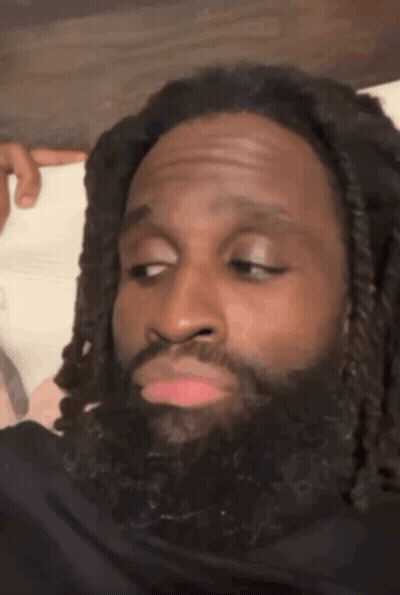 A Man With Dreadlocks And A Beard Is Making A Funny Face While Sitting On A Bed . GIF