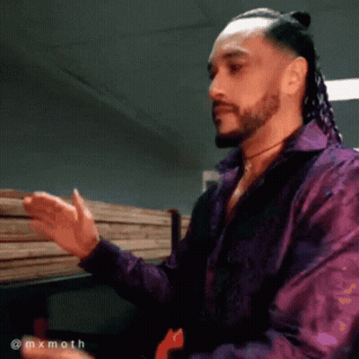 A Man With Dreadlocks And A Beard Wearing A Purple Shirt Is Standing In Front Of A Stack Of Wood GIF