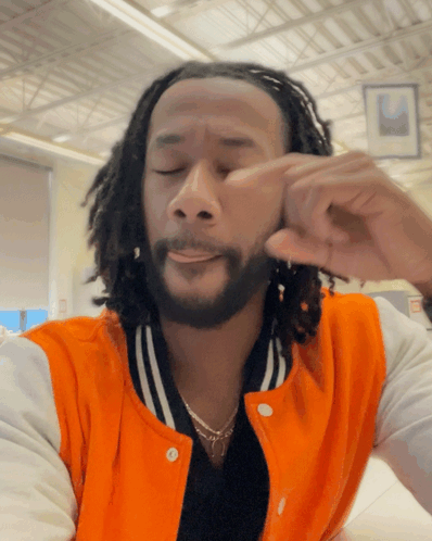 A Man With Dreadlocks And A Beard Wears An Orange Jacket GIF