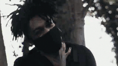 A Man With Dreadlocks And A Black Mask On His Face Is Standing In Front Of A Tree . GIF