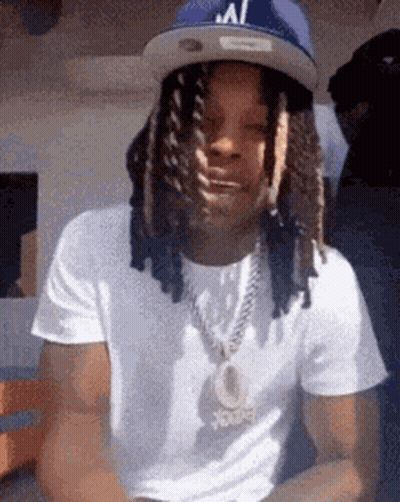 A Man With Dreadlocks And A Hat Is Wearing A White Shirt And A Chain . GIF