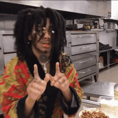 A Man With Dreadlocks And Glasses Is Giving Two Thumbs Up In A Kitchen . GIF