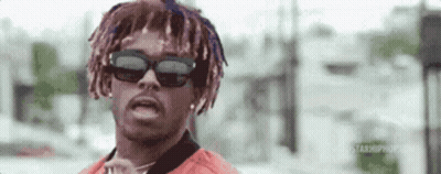 A Man With Dreadlocks And Sunglasses Is Standing In Front Of A Building And Looking At The Camera . GIF