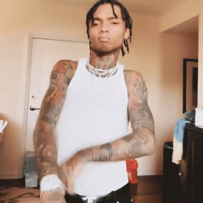 A Man With Dreadlocks And Tattoos Is Wearing A White Tank Top And Black Pants . GIF
