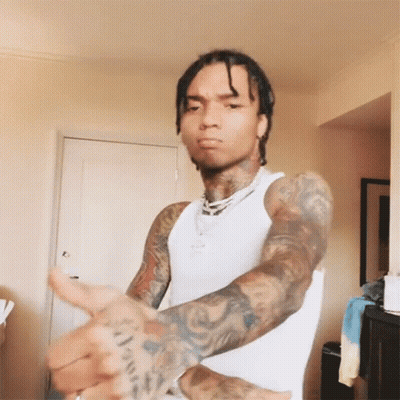 A Man With Dreadlocks And Tattoos Is Wearing A White Tank Top And Making A Funny Face . GIF