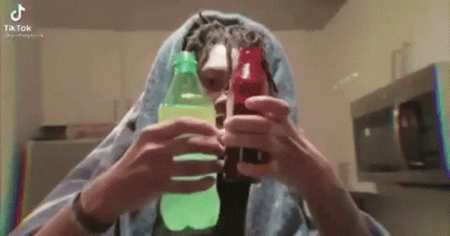 A Man With Dreadlocks Is Holding Two Bottles Of Soda In His Hands . GIF