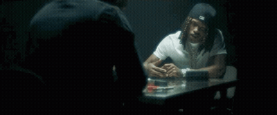 A Man With Dreadlocks Is Sitting At A Table Talking To Another Man In A Dark Room . GIF