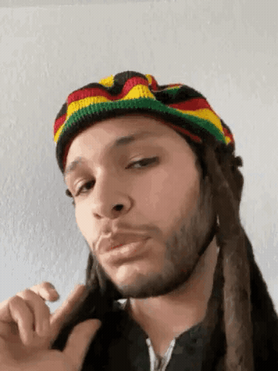 A Man With Dreadlocks Is Wearing A Rasta Hat And Making A Funny Face . GIF