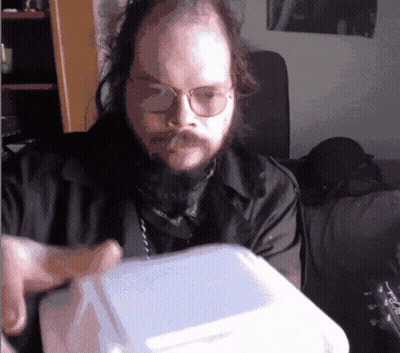A Man With Glasses And A Beard Is Holding A Styrofoam Box GIF