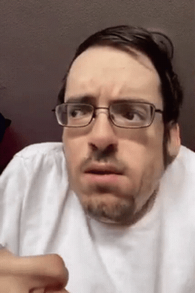 A Man With Glasses And A Beard Is Making A Funny Face While Wearing A White Shirt . GIF