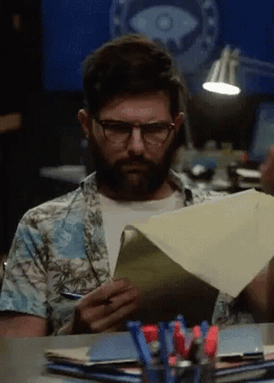 A Man With Glasses And A Beard Is Sitting At A Desk Reading A Piece Of Paper GIF