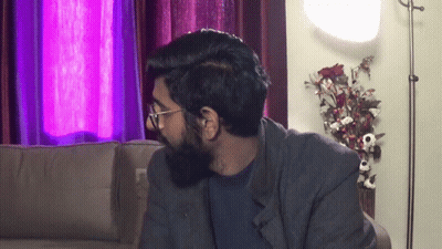 A Man With Glasses And A Beard Is Sitting On A Couch In A Living Room GIF