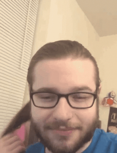 A Man With Glasses And A Beard Is Smiling While A Woman Holds His Hair . GIF