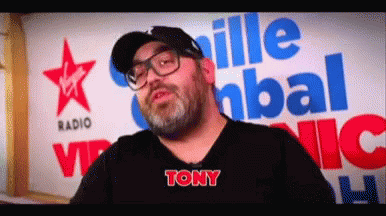 A Man With Glasses And A Beard Is Standing In Front Of A Virgin Radio Sign GIF