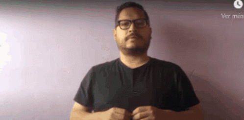 A Man With Glasses And A Beard Is Wearing A Black Shirt Against A Purple Wall . GIF