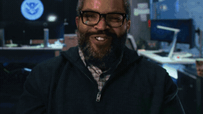 A Man With Glasses And A Beard Smiles In Front Of A U.s. Department Of State Logo GIF