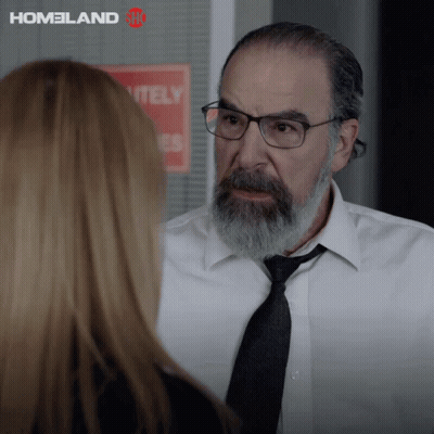 A Man With Glasses And A Beard Talks To A Woman In Front Of A Sign That Says " Homeland " GIF