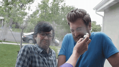 A Man With Glasses And A Blue Shirt Looks At Another Man With A Cigarette In His Mouth GIF