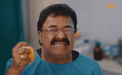 A Man With Glasses And A Mustache Is Holding An Orange In Front Of A Sign That Says Xmk GIF