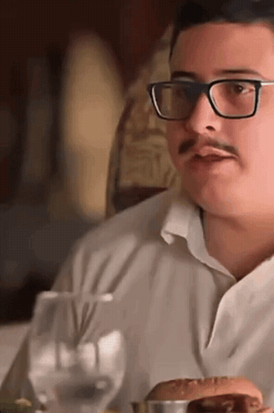 A Man With Glasses And A Mustache Is Sitting At A Table Eating A Hamburger GIF