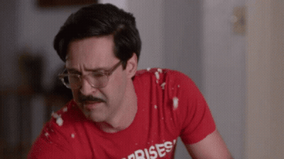 A Man With Glasses And A Mustache Is Wearing A Red Shirt That Says Surprises GIF